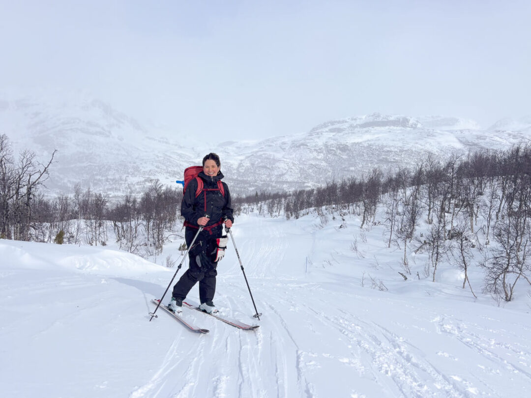 Woman ski touring in snowy Norway mountains, trekking with poles across a ski track on a winter day.