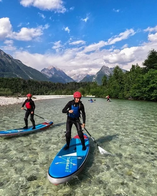 Women stand-up paddleboarding on a clear Alpine river, with mountains and forest in the background