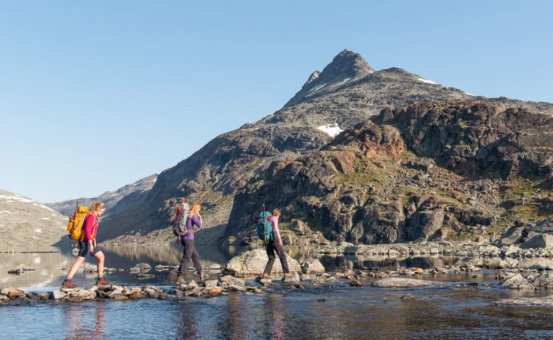 Women hiking along a mountain lake in Norway’s rugged landscape, with peaks in the background.