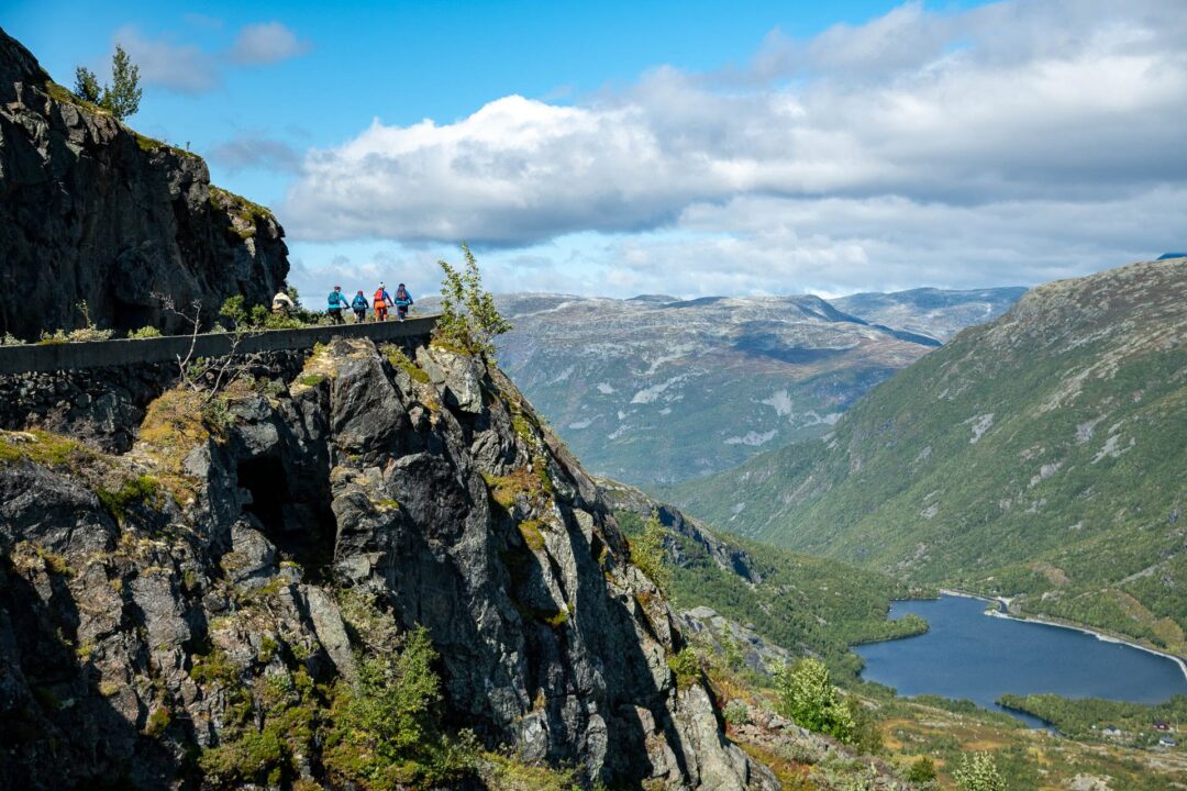 Hikers trekking along a rocky cliff above a fjord and green valley under a cloudy sky