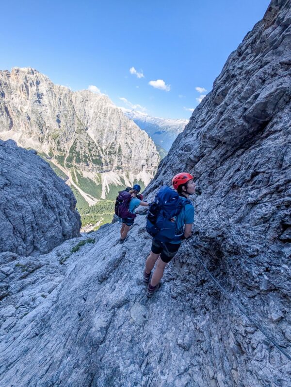 Women climbing via ferrata on rocky alpine cliffs with cables, overlooking a mountain valley in the Dolomites