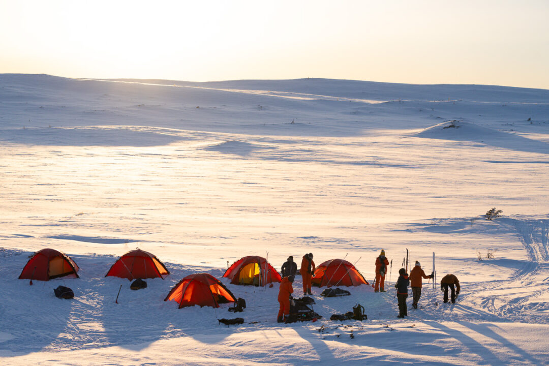 Winter camping with tents in a snowy landscape at dusk, with people and gear on the hillside