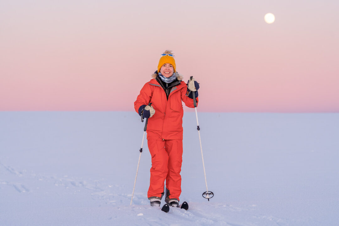 Woman skiing on a snowy landscape at dusk, wearing red winter gear and holding ski poles