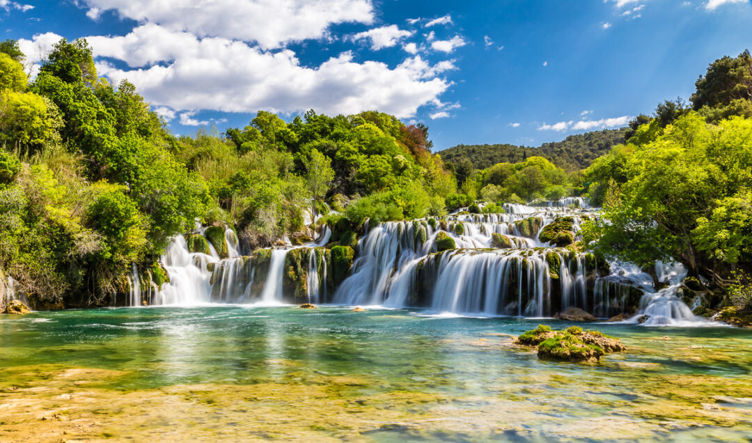 Multi-tier waterfall cascading into a turquoise river pool surrounded by green forest