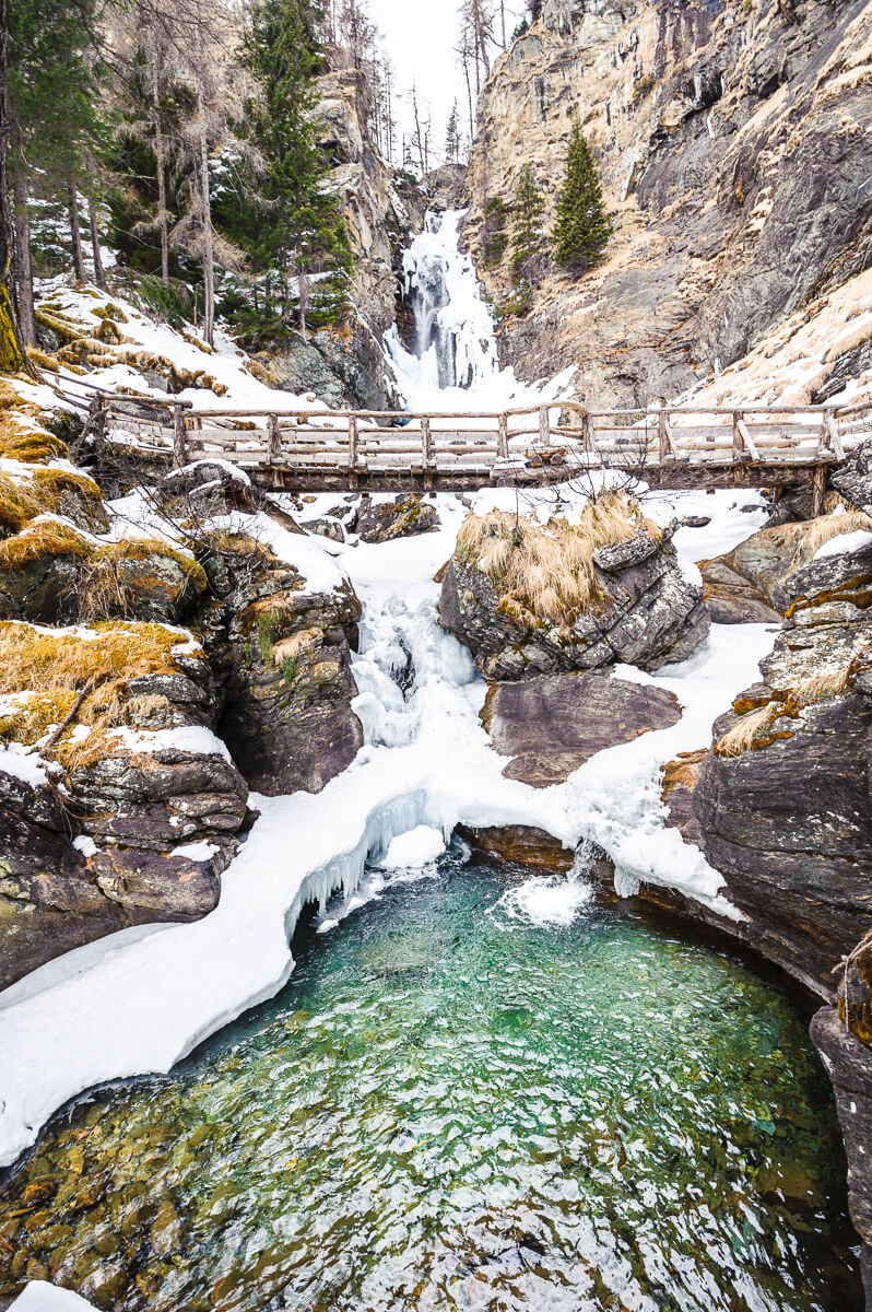 Snowy Dolomites gorge with a waterfall, wooden bridge and winter hot spring pool