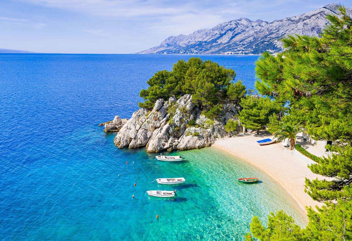 Secluded coastal beach with small boats, clear turquoise water and pine trees beside distant mountains