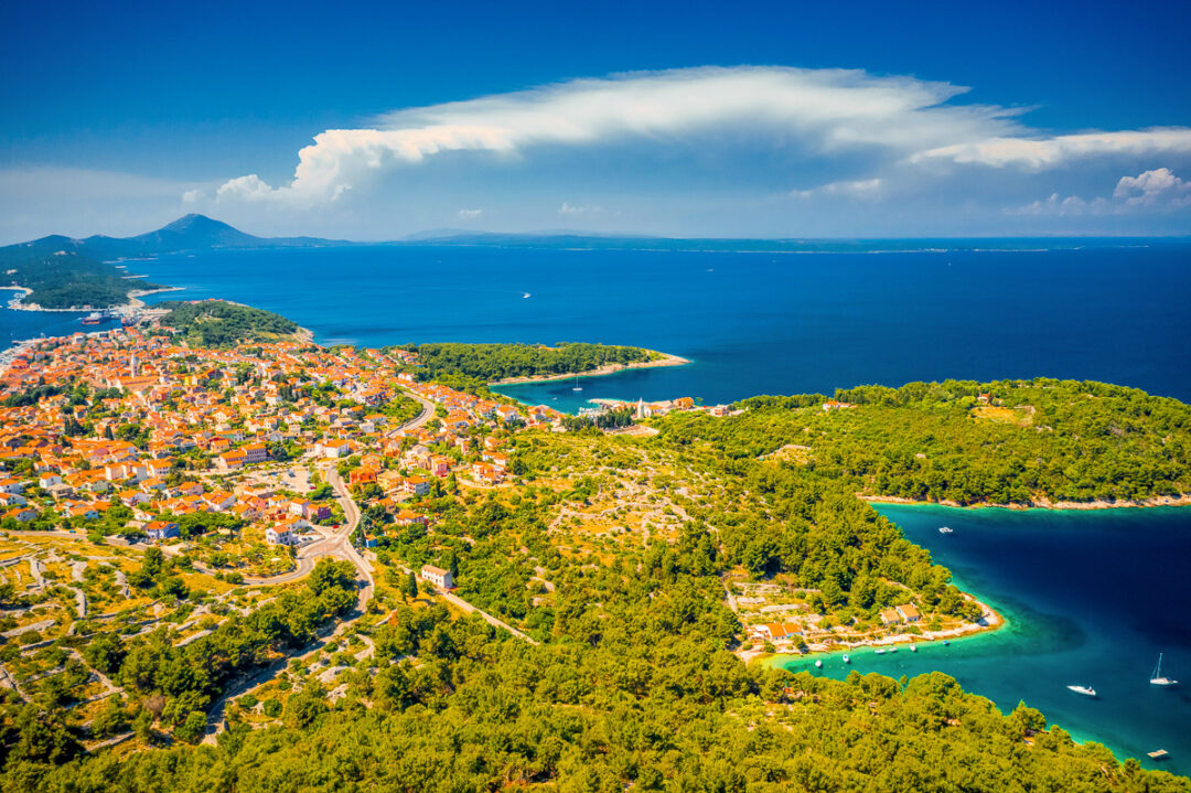 Coastal aerial view of a hillside town, sandy coves and boats along a calm blue sea