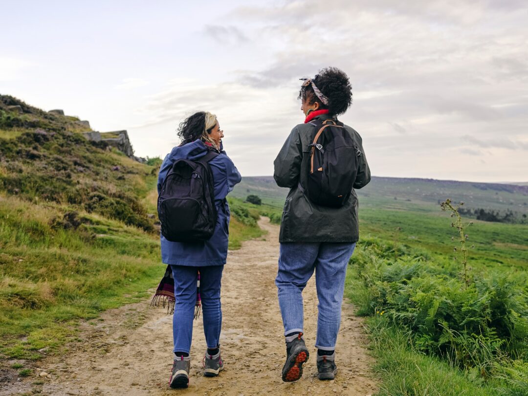 Two women hiking on a rural footpath with backpacks, overlooking grassy hills under a cloudy sky