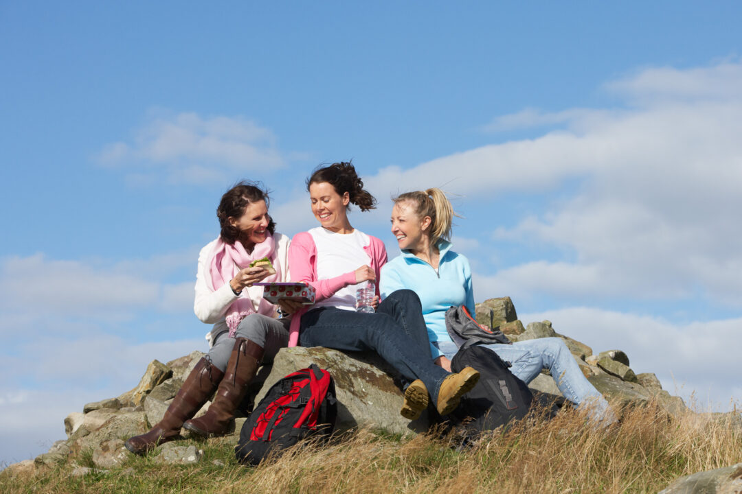 Three women on a rocky hilltop during a hiking break, with backpacks against a blue sky