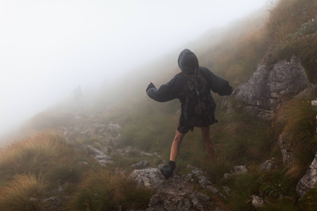 Women hiking along a rocky mountain trail in thick mist, wearing waterproof jackets on a foggy hillside