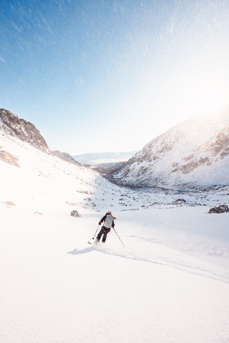 Woman skiing downhill through a snow-covered alpine valley with mountains under a clear blue sky