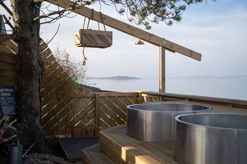 Outdoor sauna hot tub on a wooden deck overlooking the sea, with a sign and fence in the UK countryside