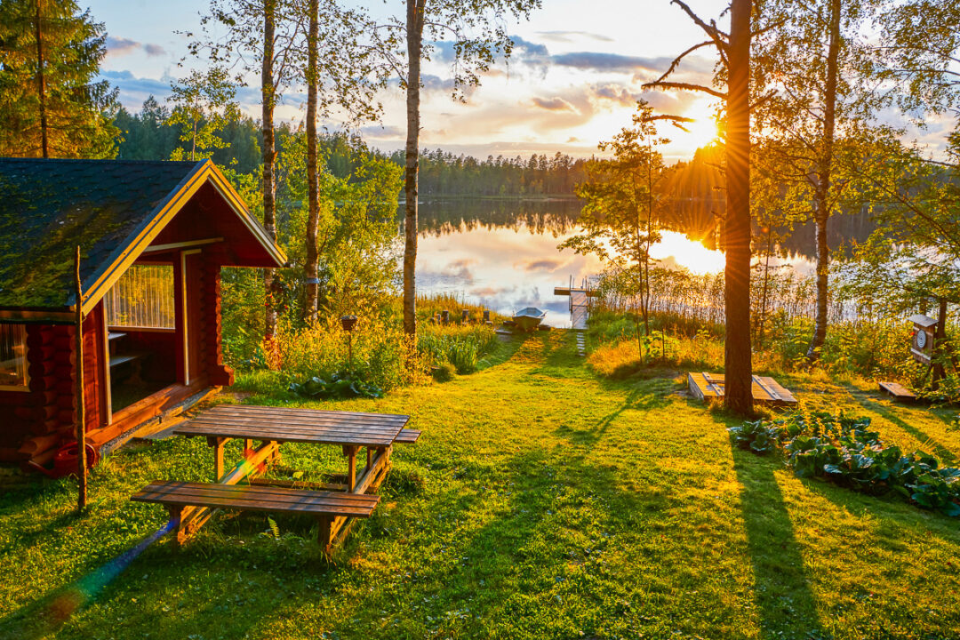 Riverside cabin and picnic bench on a grassy lawn at sunset with trees and calm water in the background