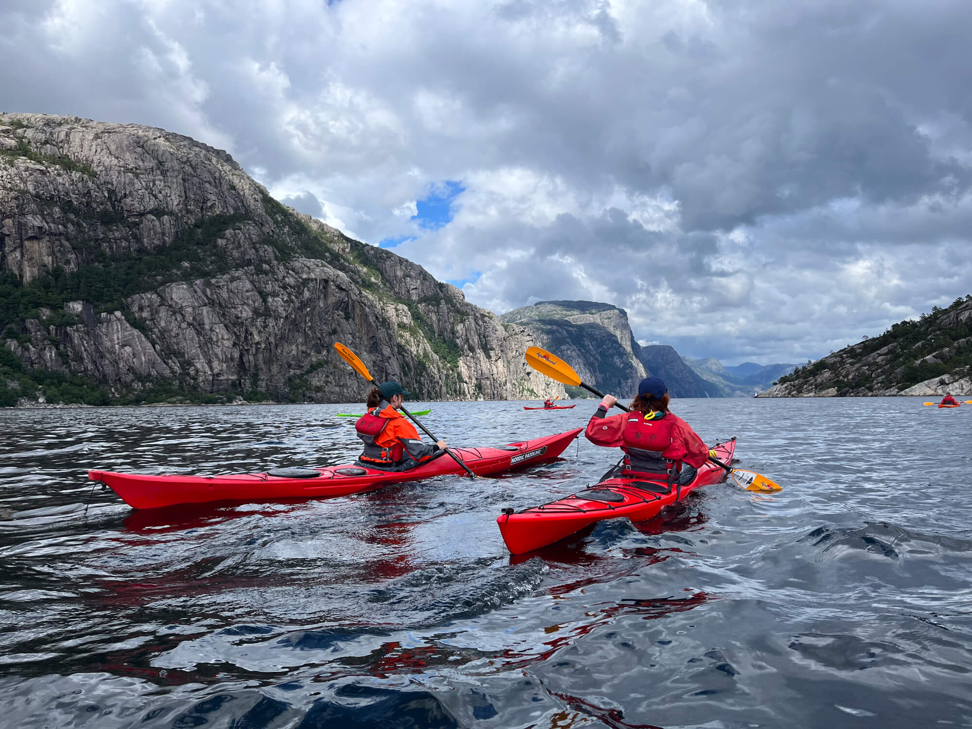 Women kayaking on a fjord in Norway, paddling past steep cliff faces under cloudy skies