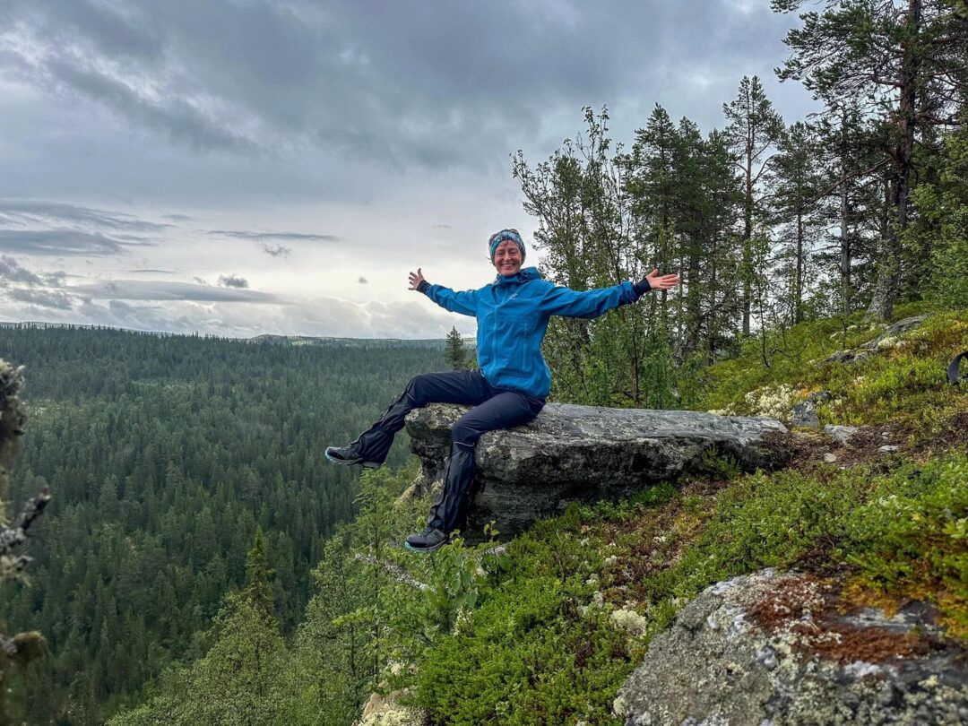 Woman on a rock during hiking in a forested mountain valley, wearing a blue outdoor jacket outdoors.