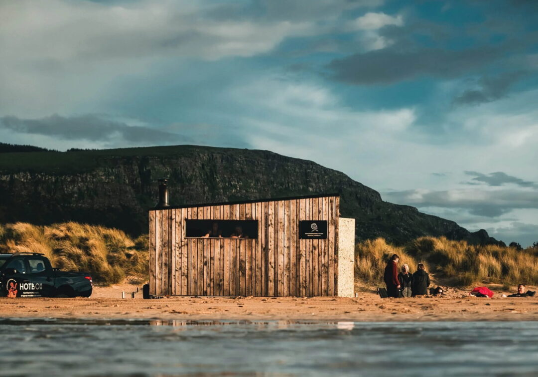 Outdoor wooden sauna hut by a UK beach, with people outside near dunes and mountains