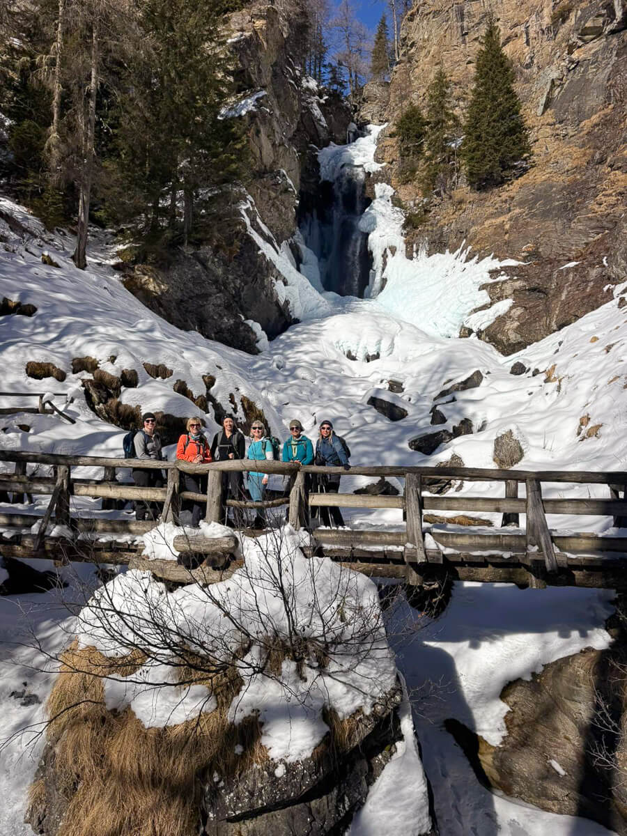 Women in winter hiking gear on a wooden bridge beside a snowy mountain waterfall in the Alps