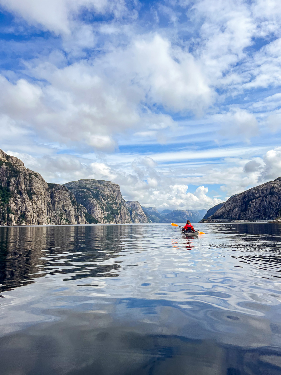 Woman kayaking on calm fjord water between steep mountains under a blue sky