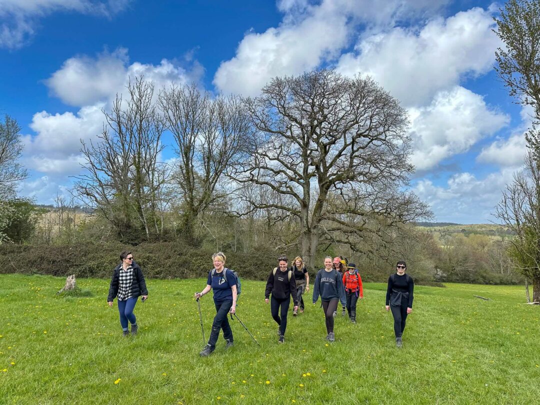 Group of women on a guided day hike, walking across a grassy field under a blue sky with trees
