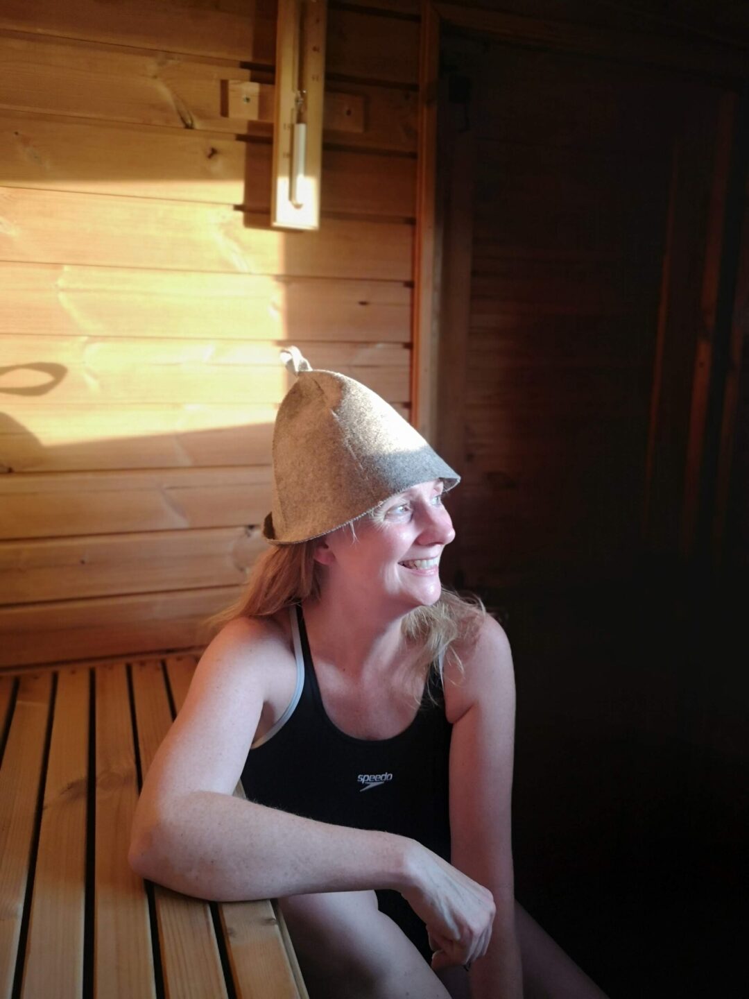 Woman relaxing in a wooden sauna, wearing a sauna hat and smiling.
