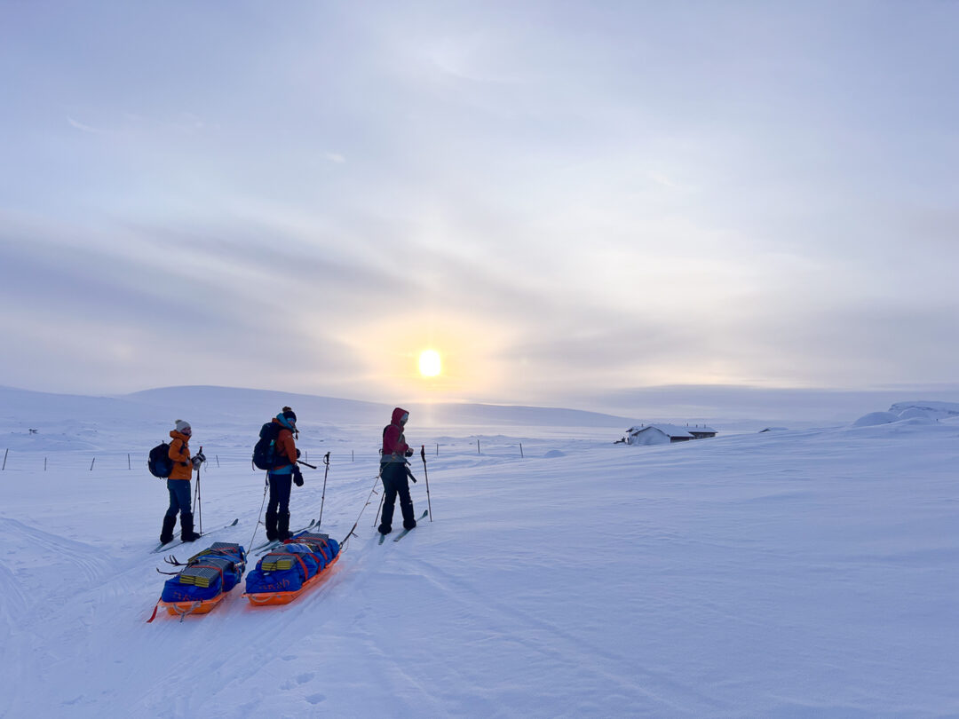 Women on skis trekking snowy mountains at sunrise with toboggans, distant hills and ski track in view