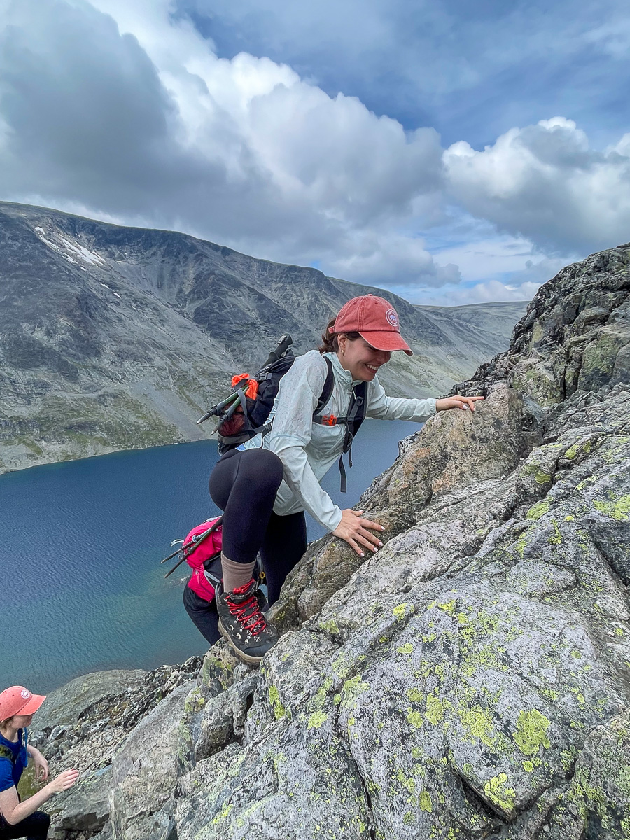 Woman hiking on a rocky mountain ridge above a blue alpine lake, with other women on adventure holiday in Europe