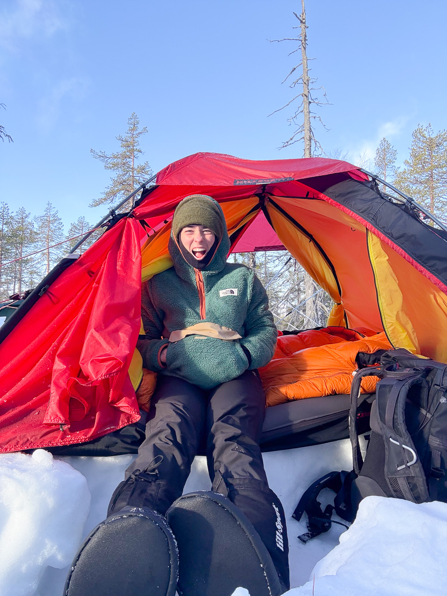 Women winter camping in a snowy forest, sitting inside a brightly coloured tent outdoors.