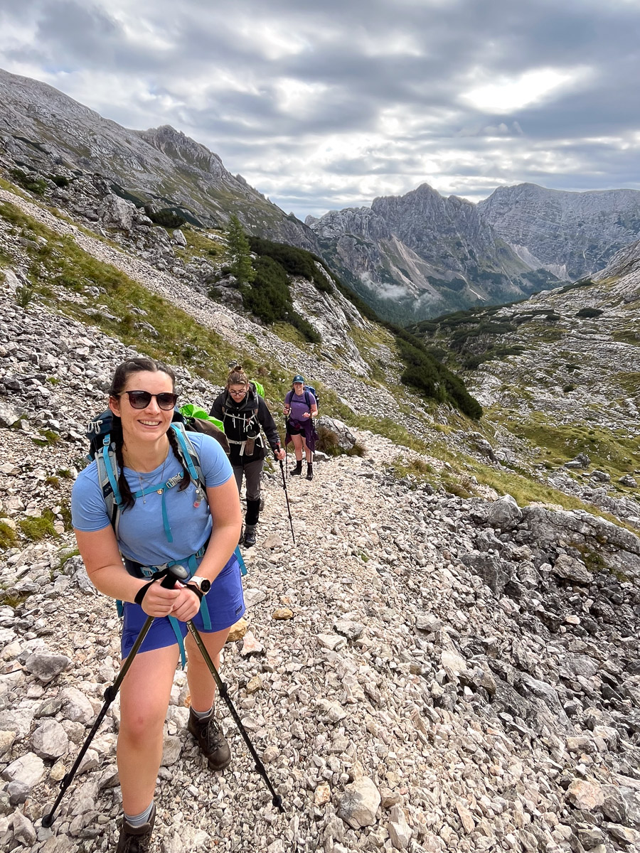 Women hiking on a rocky mountain trail under a cloudy sky, with alpine valley views in the background