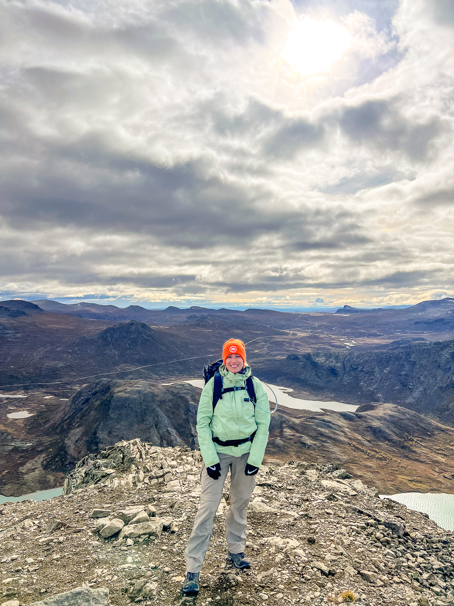 Woman hiking on a mountain summit with a scenic valley and lake below under cloudy skies