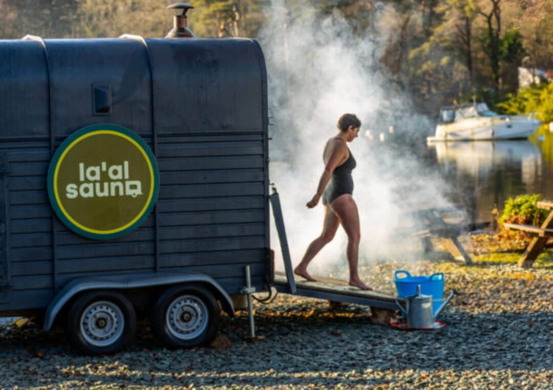 Woman using a barrel sauna, steaming outdoors by a “la’gal sauna” sign in the UK countryside