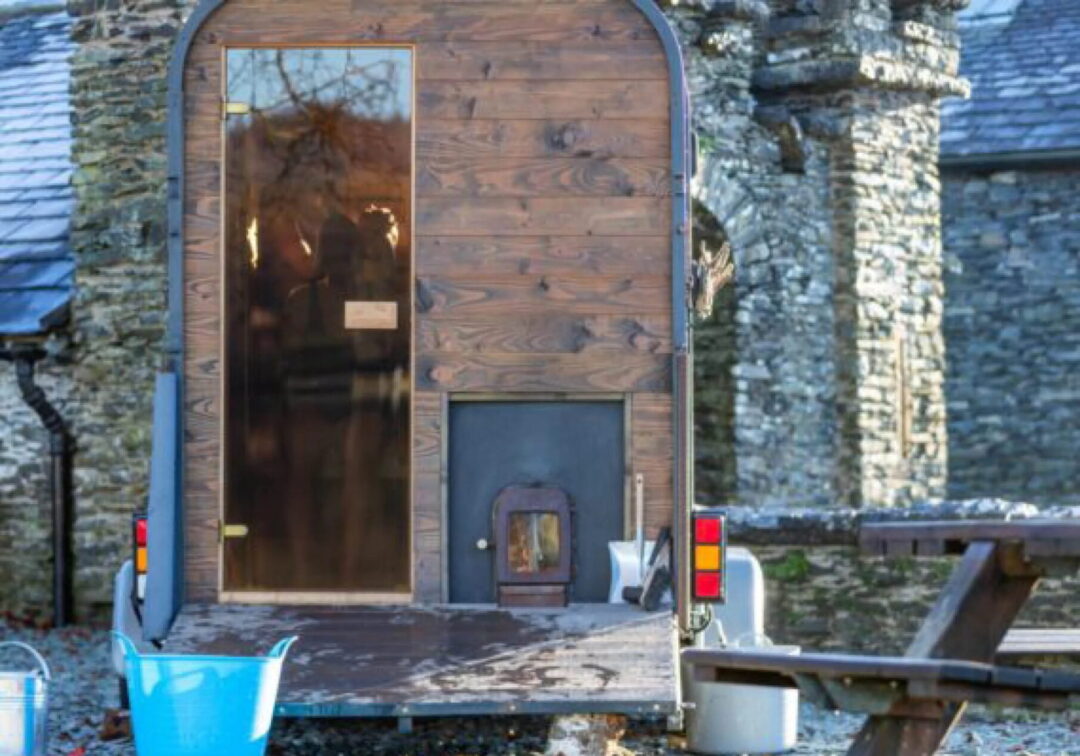 Outdoor sauna entrance with wooden door set in stone building in the UK, with bench and wheelbarrow nearby
