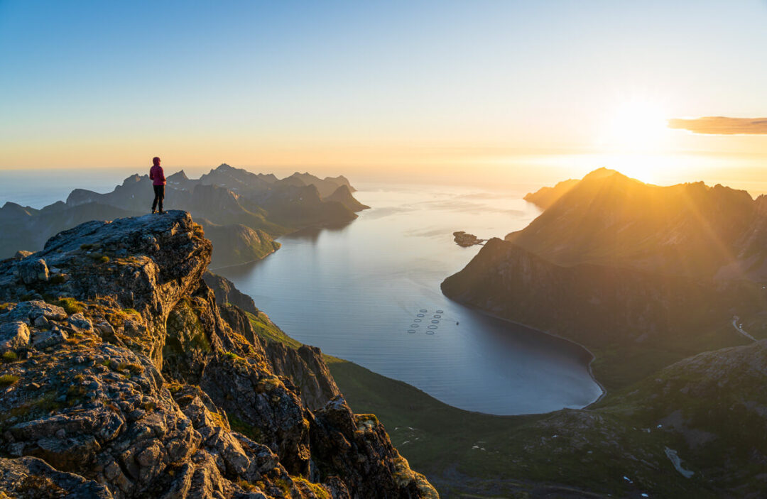 Woman stands on a rocky cliff at sunrise, overlooking a fjord landscape
