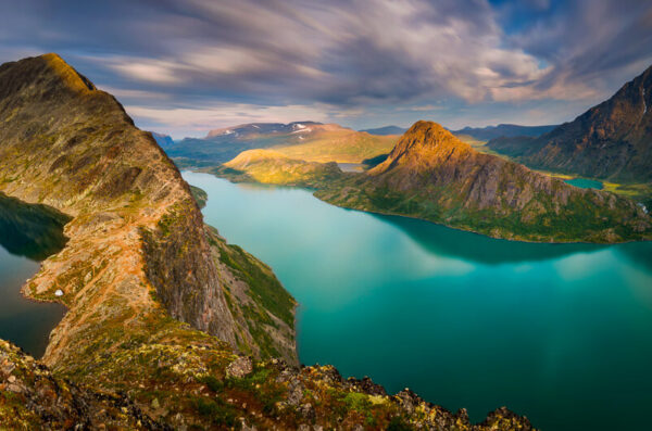 View from a mountain ridge over a turquoise fjord in Norway, with surrounding peaks and valleys at sunset.