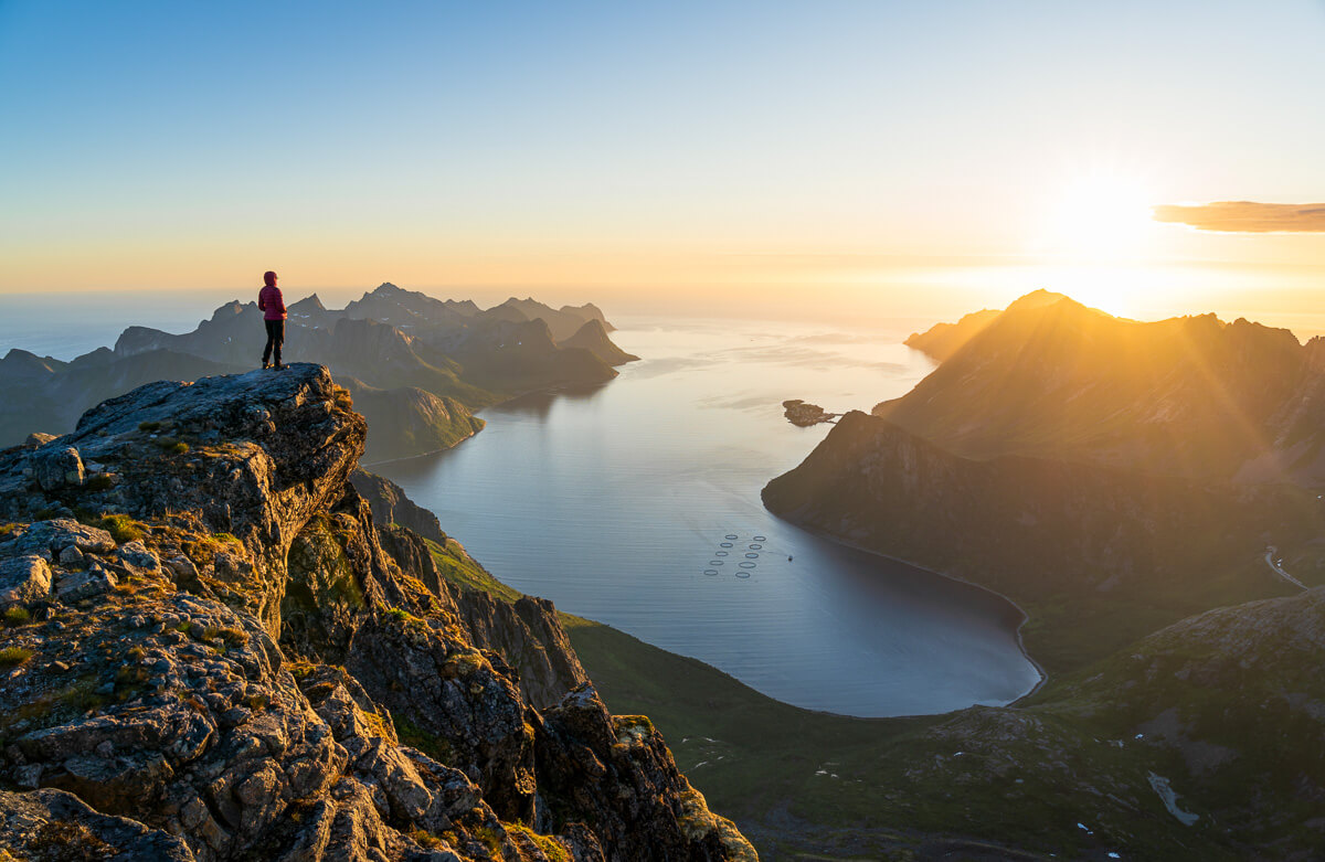 Woman stands on a rocky cliff at sunrise, overlooking a fjord landscape