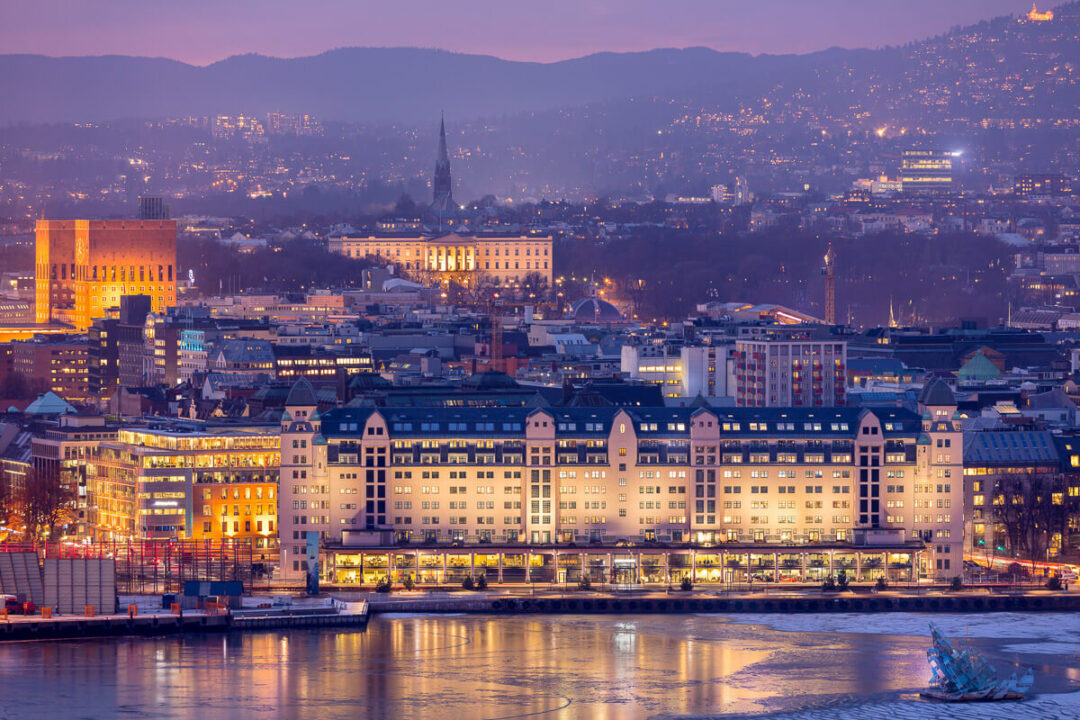 Evening skyline over a river in a European city, with illuminated buildings and church spires in the distance