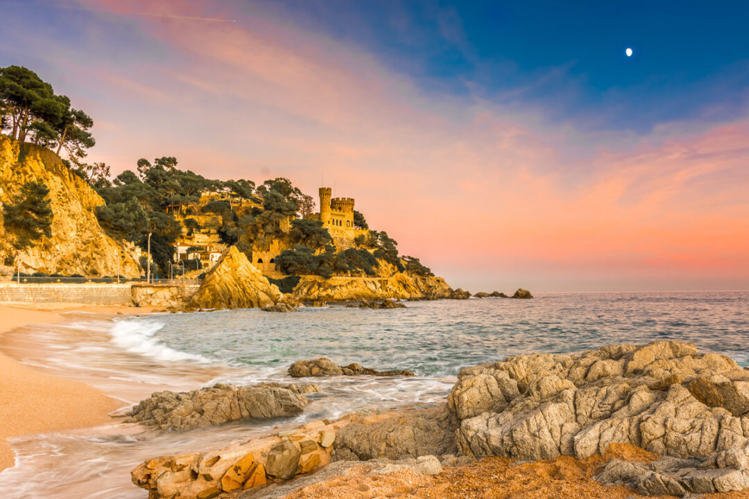Rocky beach and lighthouse on a Mediterranean coastline at sunset with calm waves and orange sky