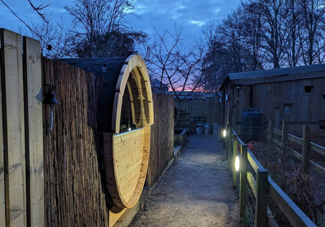 Outdoor wooden barrel sauna in a fenced yard at dusk, with warm light from the doorway.