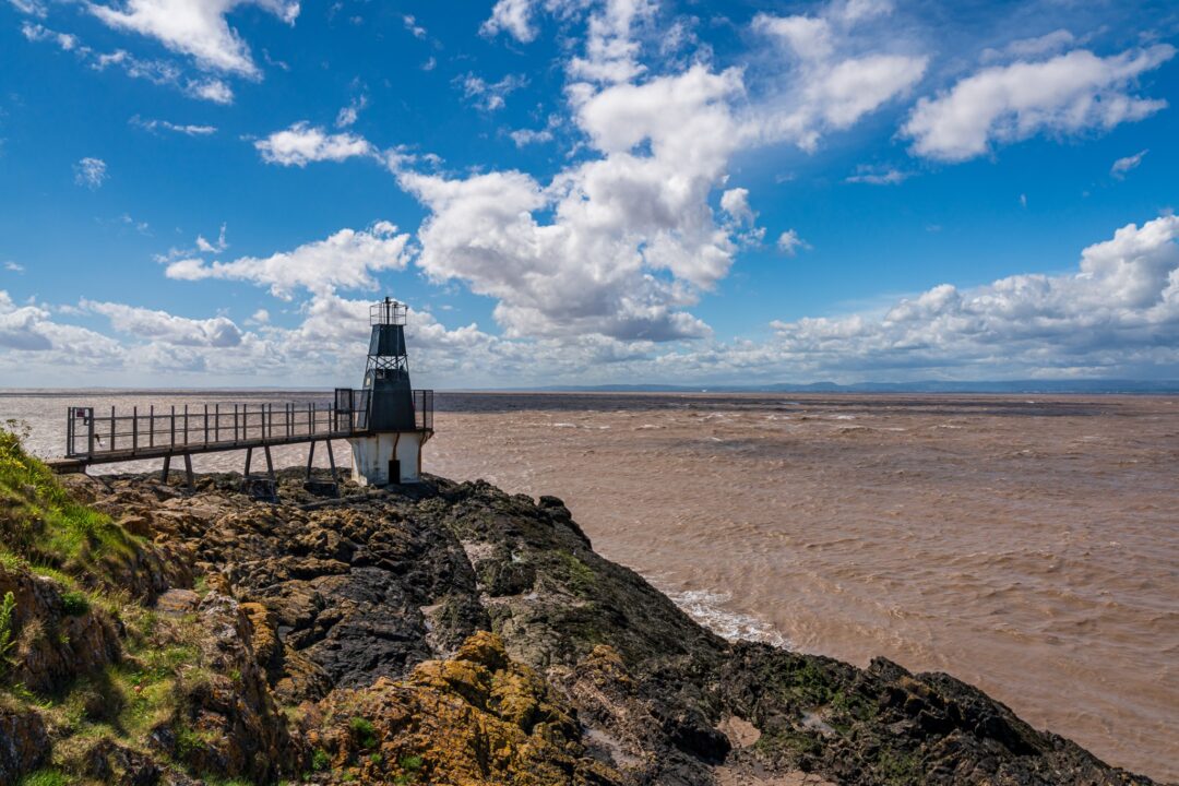 Coastal viewpoint on rocky shoreline with sea and lighthouse, under blue sky and clouds near Bristol