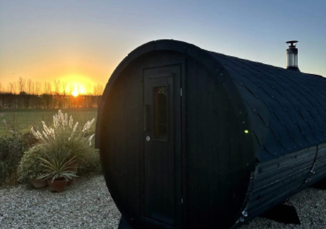 Black barrel sauna in a countryside garden at sunset, with rural trees and gravel path in the UK.