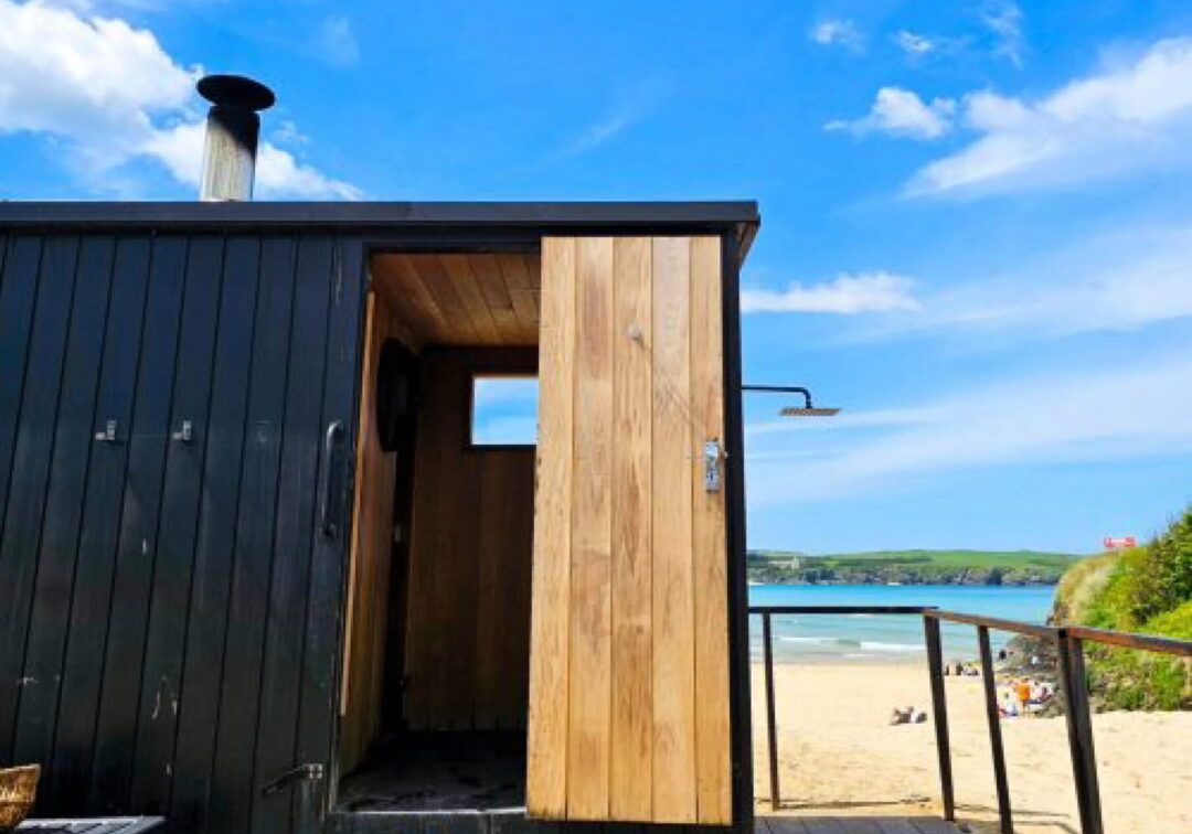 Sauna cabin doorway opening onto a beach and sea under a blue sky