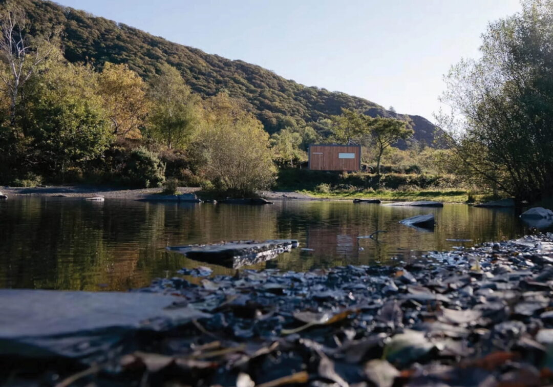 Riverbank with rocks and calm water, wooded hills in the background, and a small wooden sign by the shore.