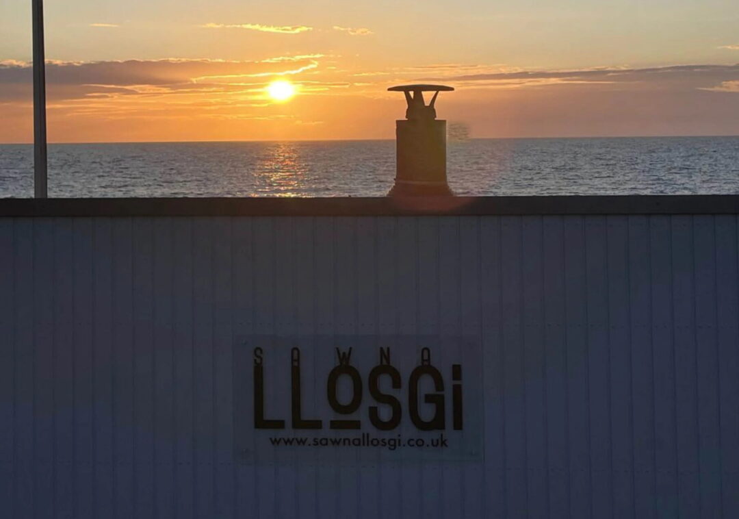 Lösgi sauna sign with URL “www.sawnalosgi.co.uk” by the sea at sunset, UK