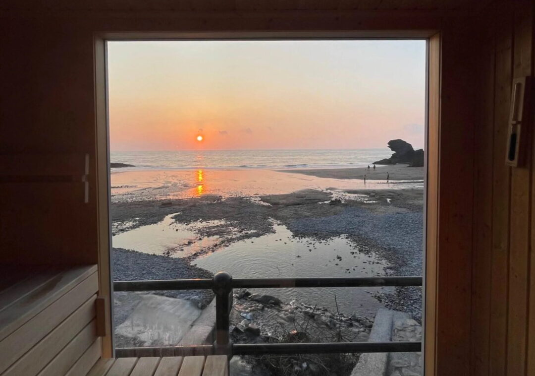 Sauna window view of a beach at sunset, with sea waves and sand visible through the open doorway