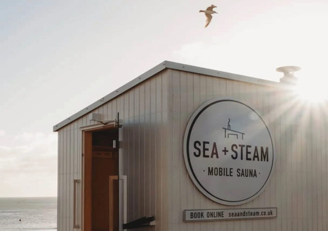 Mobile sauna hut with “SEA + STEAM” sign, by the sea in the UK at sunset