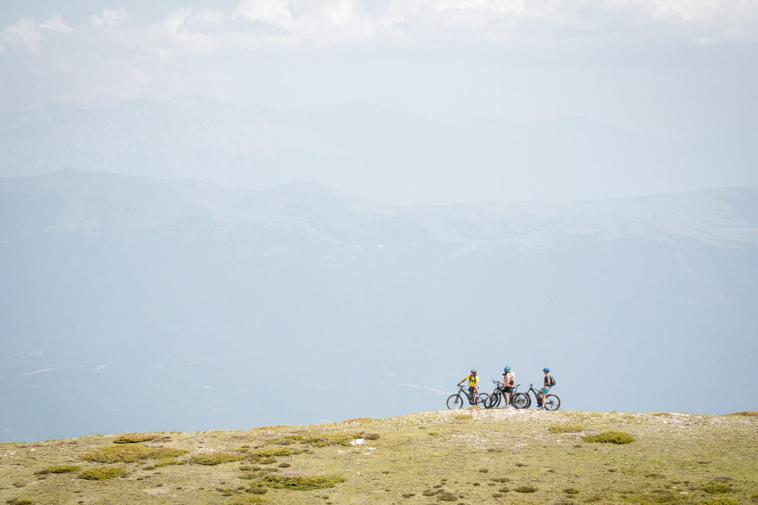 Hikers trek across a grassy mountain ridge with backpacks, overlooking misty peaks in the distance