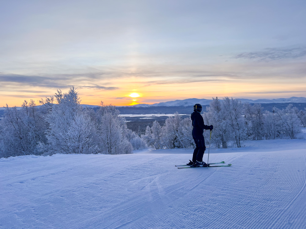 Woman snowboarding on a snowy piste at sunrise, with frosted trees and distant hills in winter landscape