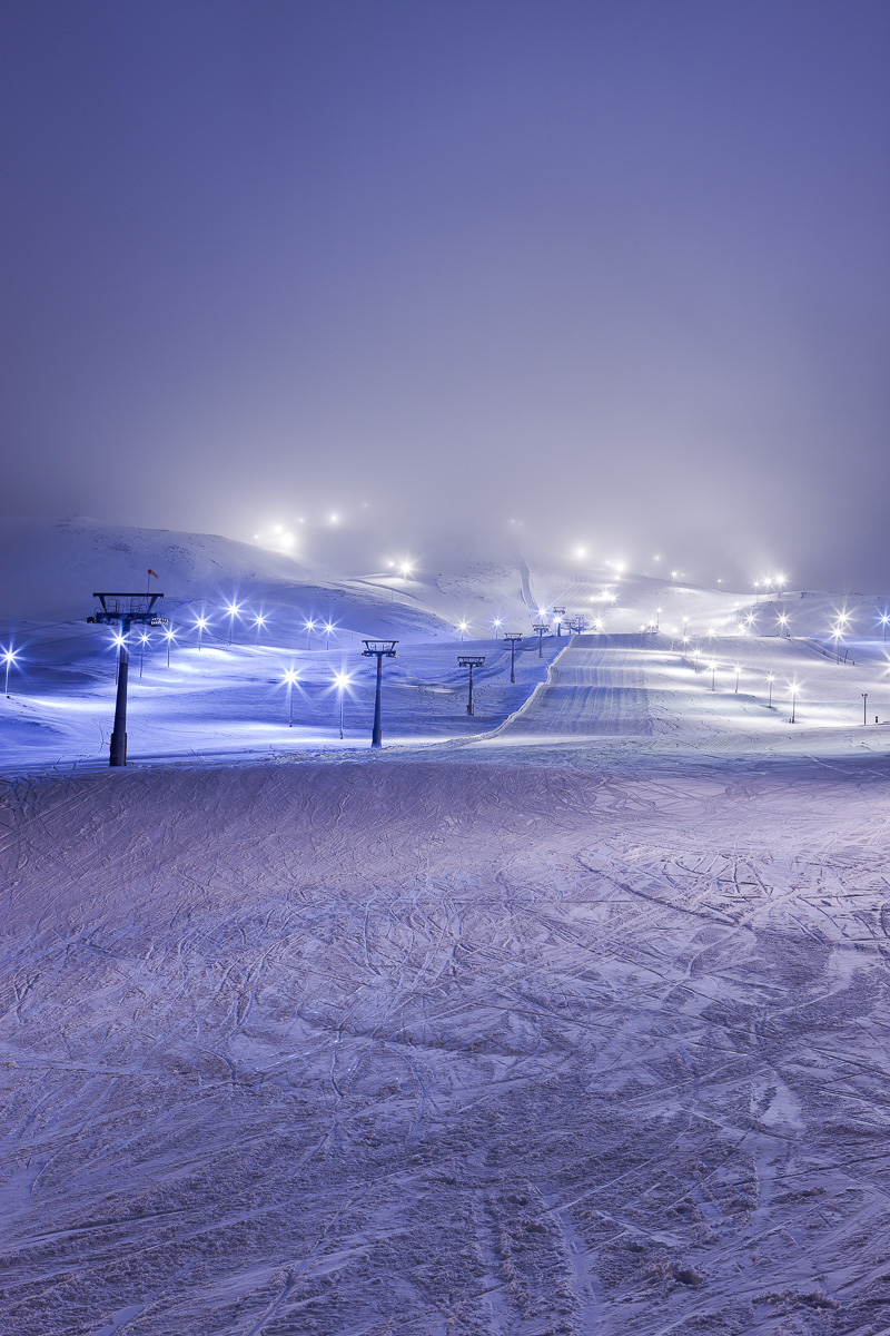 Snowy ski slope at night with illuminated ski lift stations and lights across the resort