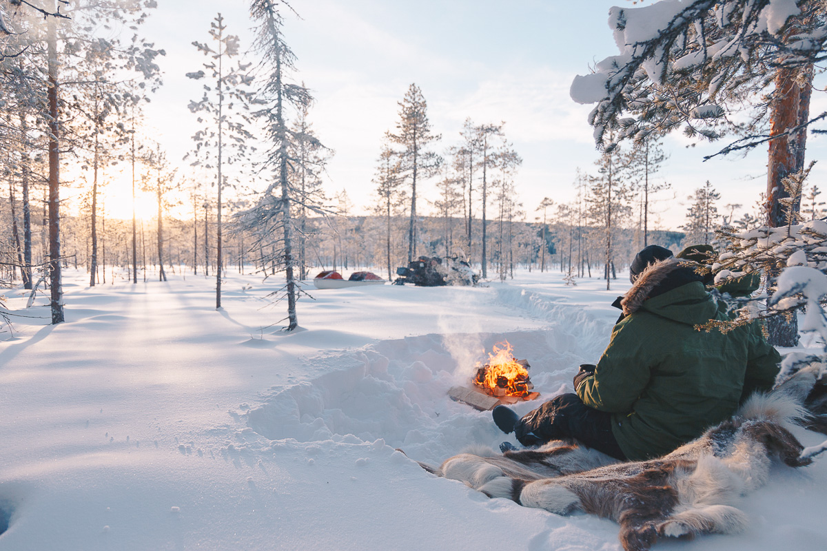 Women camping in a snowy forest, warming hands by a small campfire in winter landscape
