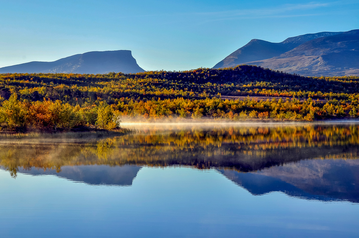 Mountain lake landscape with autumn trees and reflections, blue sky over distant hills