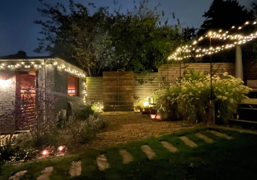 Outdoor sauna in a garden at night, lit with string lights and surrounded by shrubs.